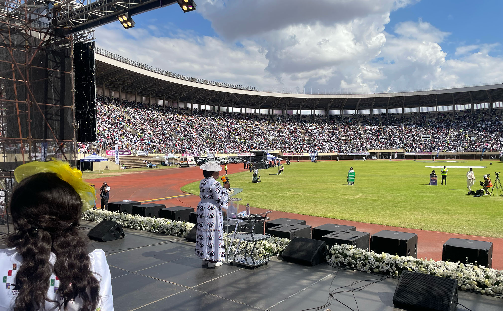 A wide shot showing Apostle Eunor Guti on stage addressing a large crowd at the Zimbabwe National Sports stadium, filled with members of ZAOGA FIFMI who had come to celebrate the life of Archbishop Ezekiel Guti
